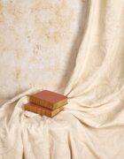 A swathe of cream on cream fabric draped in front of an antique stone wall with old books supporting the fabric.